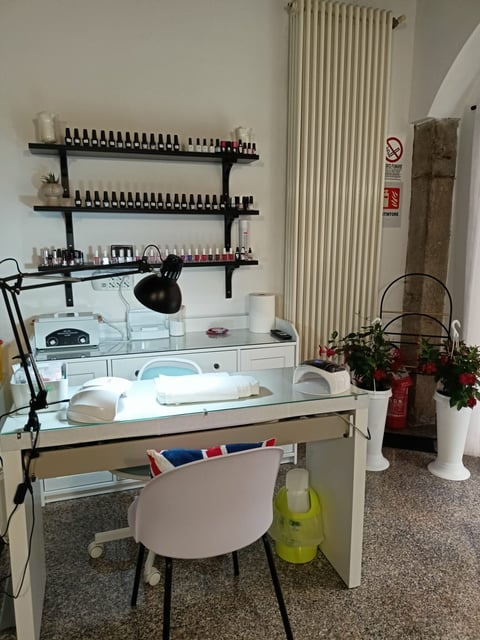 A nail salon workstation with glass desk, white chair, adjustable lamp, nail polish shelving, and radiator heater in background